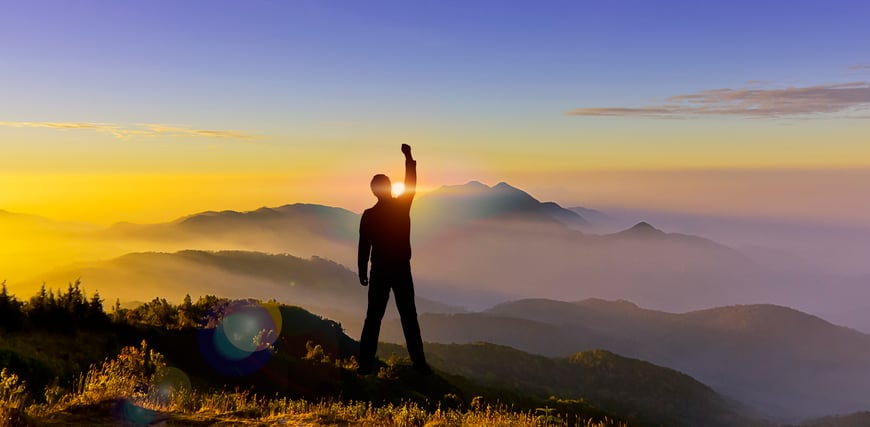 This image shows a man standing atop a height, silhouetted against the background of a setting sun with mountains in the distance and mists in the regions below. His fist is raised, showing his triumphal mood as he continues on his quest to transform his health. 