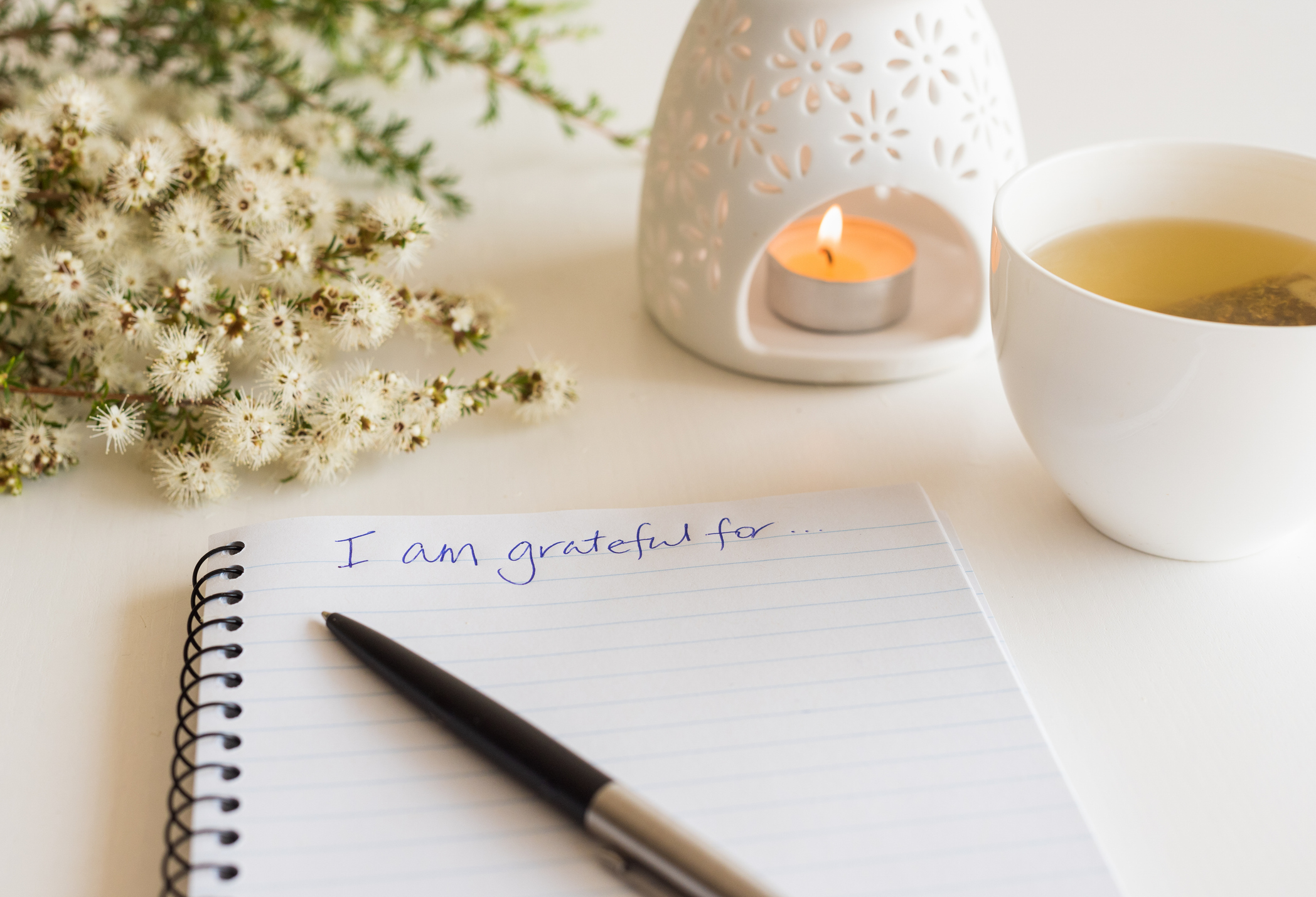 This image depicts the first steps of writing a gratitude list from a first-person perspective. There is an open notebook with the words "I'm grateful for" spelled out in the top margin. There is a candle and some small flowers arrayed on the desk as well. 
