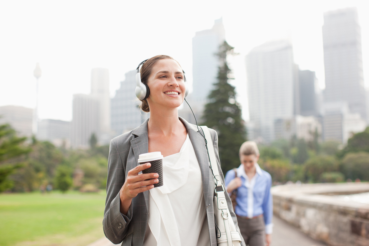 A smiling woman gets some exercise on her daily walk to work. She is holding a beverage in her hand, and there is a city skyline in the background.