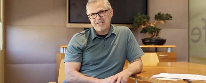 Man with gray hair and glasses sits at a round wooden conference table in an office, hands folded, with a TV screen and plant behind him.