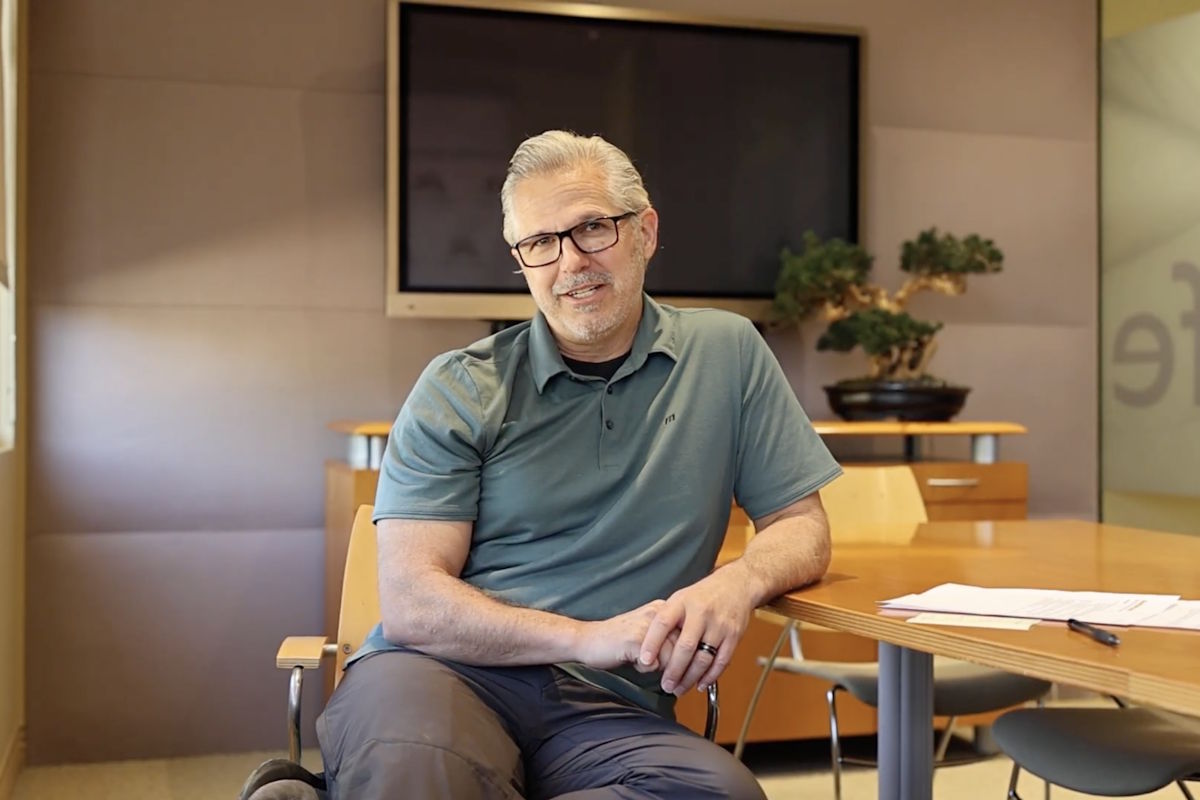 Man with gray hair and glasses sits at a round wooden conference table in an office, hands folded, with a TV screen and plant behind him.