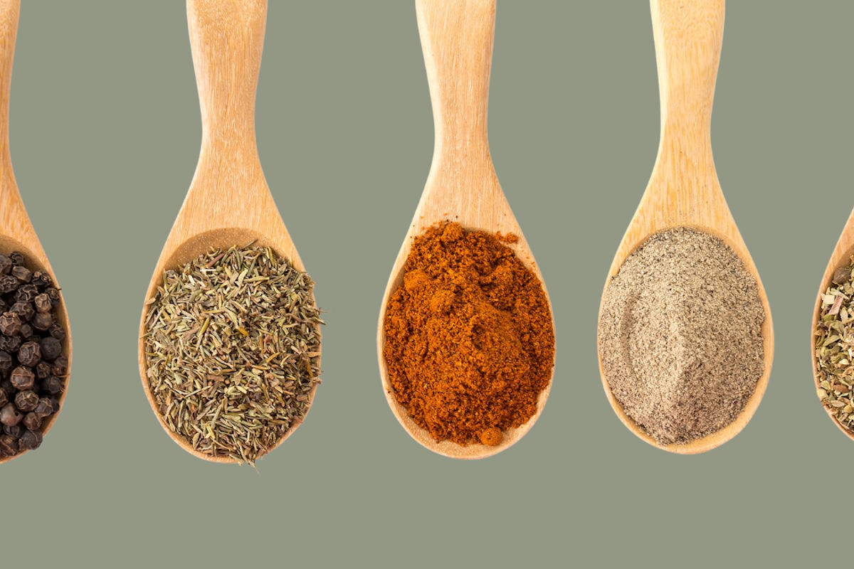 Row of five wooden spoons, each with a different spice: peppercorns, dried herbs, paprika, ground pepper, and herb mix.