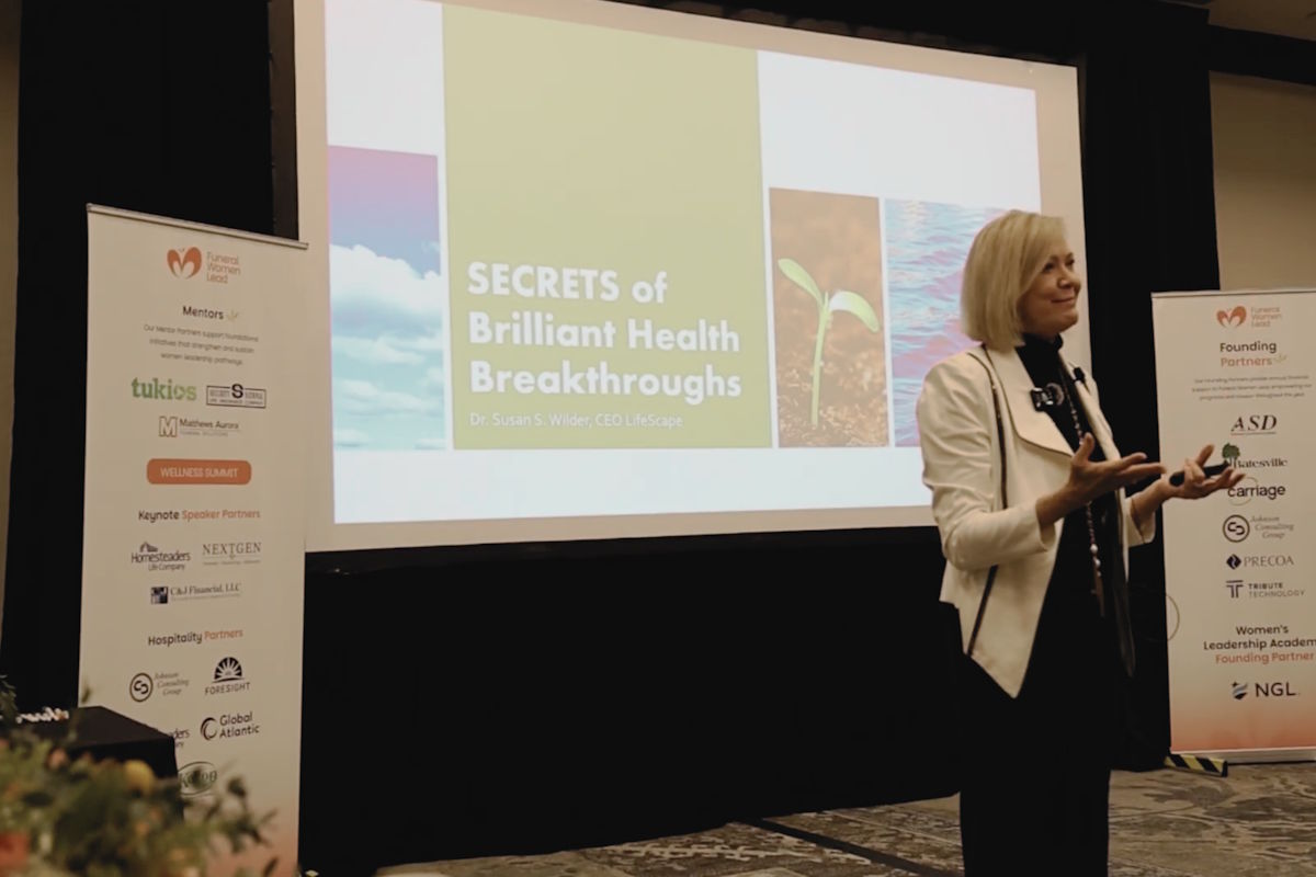 Female presenter on stage gesturing while slide reads 'Secrets of Brilliant Health Breakthroughs'.