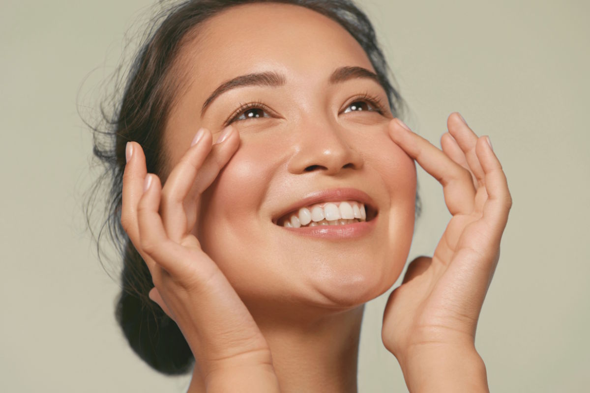 Smiling woman gently cupping her cheeks with both hands, looking upward.