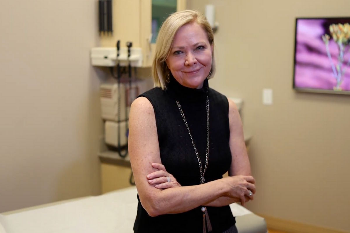 A smiling woman in a sleeveless black top stands with arms crossed in a medical exam room.