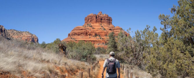 Hiker with backpack walking orange dirt trail toward red rock formation under a clear blue sky.