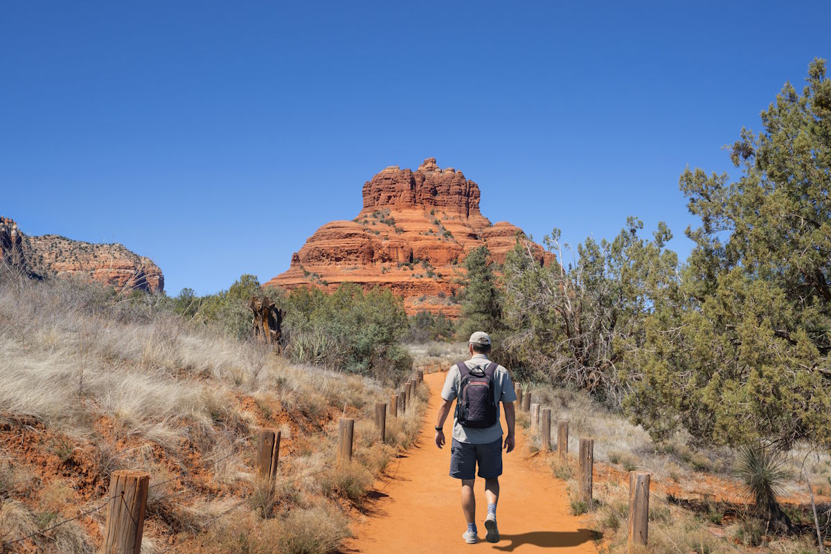 Hiker with backpack walking orange dirt trail toward red rock formation under a clear blue sky.