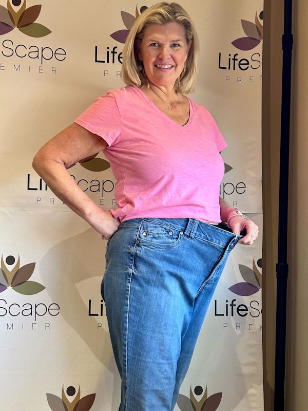 Smiling woman in a pink t-shirt holds out loose jeans at the waist, showing weight loss; backdrop features LifeScape Premier logos.