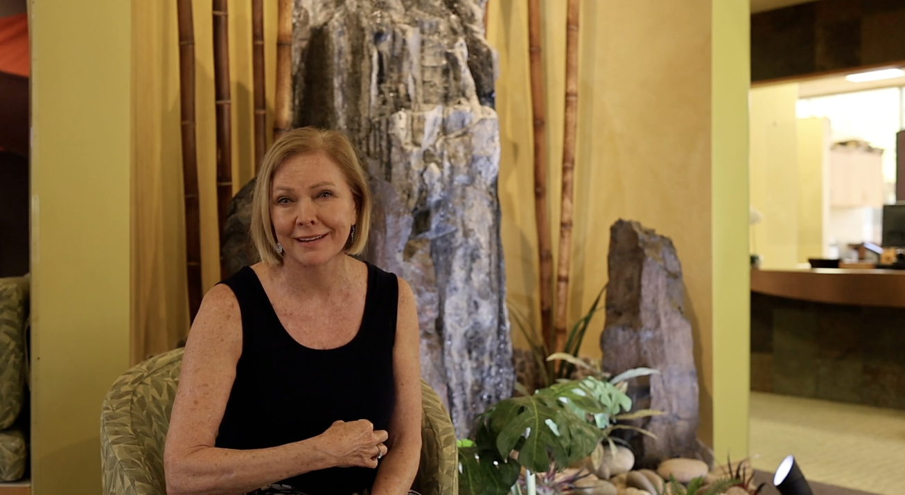Dr Susan Wilder | LifeScape Goal of Functional Medicine Blog Image - LifeScape Concierge Primary Care, Aesthetics & Nutrition in Scottsdale Smiling woman in a black sleeveless top seated in a lobby with a rock feature and plants behind her.