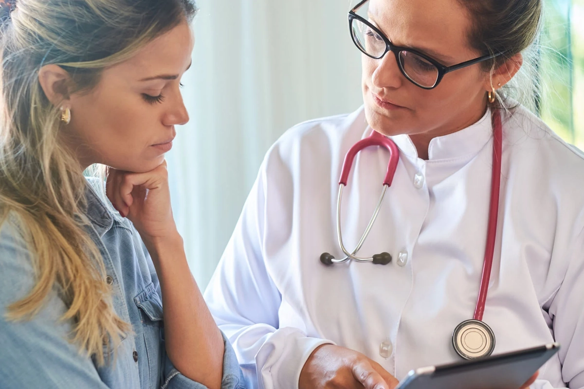 Doctor in white coat with stethoscope consults a patient, both looking at a tablet or clipboard together in a clinic.