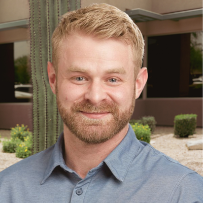 LS Lucas Walk Headshot - LifeScape Concierge Primary Care, Aesthetics & Nutrition in Scottsdale Portrait of a smiling man with short blond hair and a beard, wearing a blue button-down shirt, outdoors near a cactus and building landscaping.