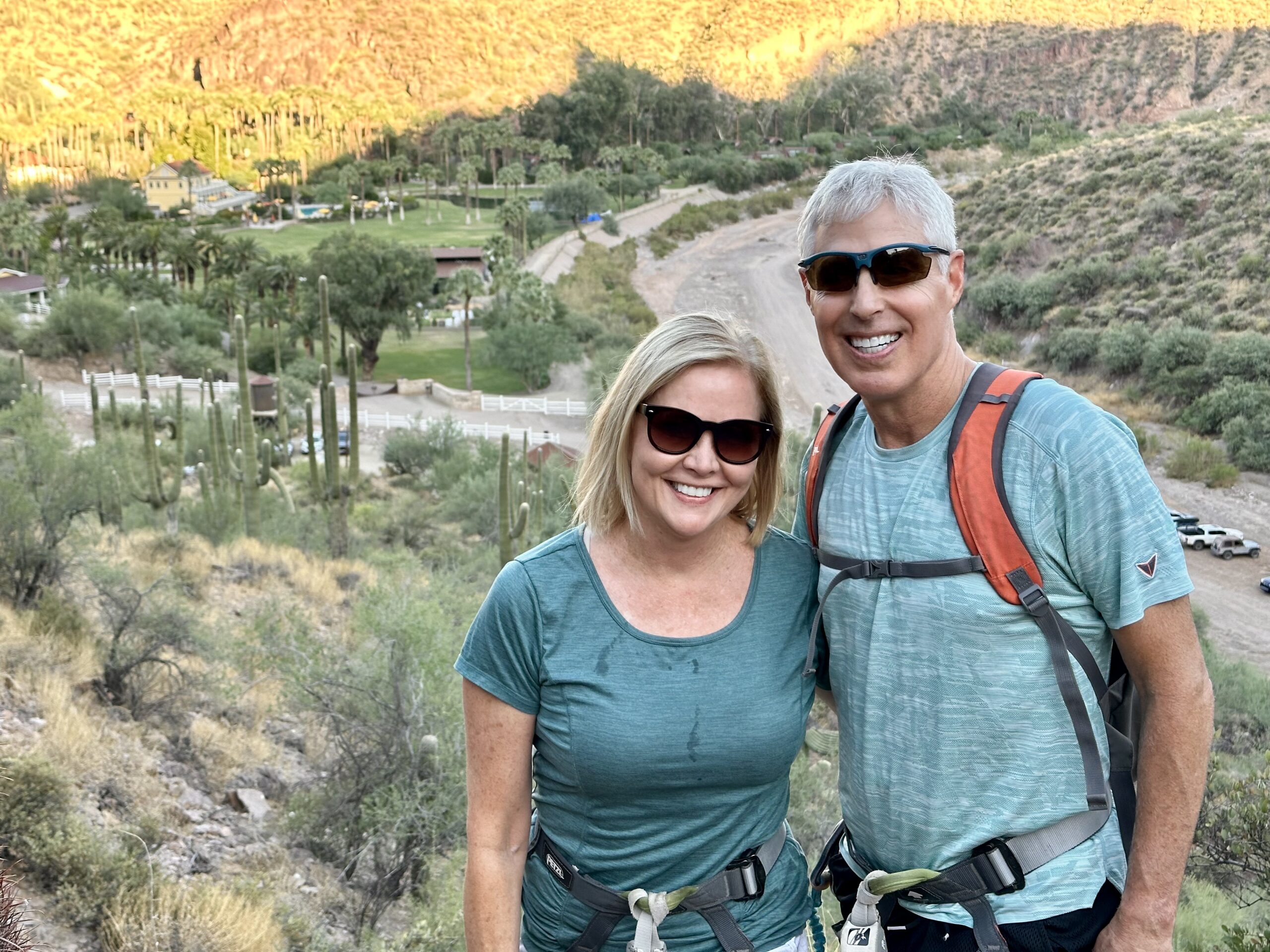 Smiling couple in teal shirts posing on a rocky trail with a desert valley, palm trees, and yellow cliffs in the background, wearing hiking backpacks and sunglasses.